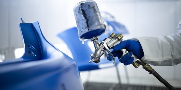Car painter in protective clothes and mask painting automobile bumper with metallic paint and varnish in chamber workshop.